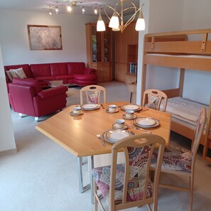 A dining area with a wooden table set for four, surrounded by patterned chairs. A red sofa and a bunk bed are also visible in the room. - Apartment '3' with Balcony, Shared Garden and Wi-Fi (Goslar)