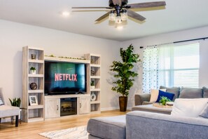 Ein Wohnzimmer mit einem Flachbildfernseher, der 'NETFLIX' anzeigt, einem Deckenventilator, einer Topfpflanze und einem grauen Sofa.