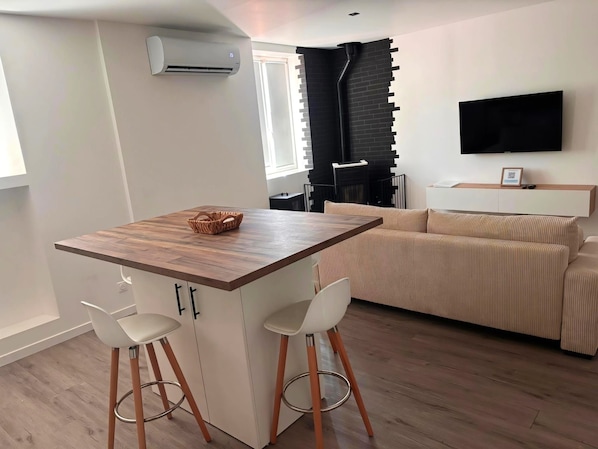 A modern kitchen with a wooden countertop, two white bar stools, a black fireplace, a beige sofa, and a flat-screen TV mounted on the wall.