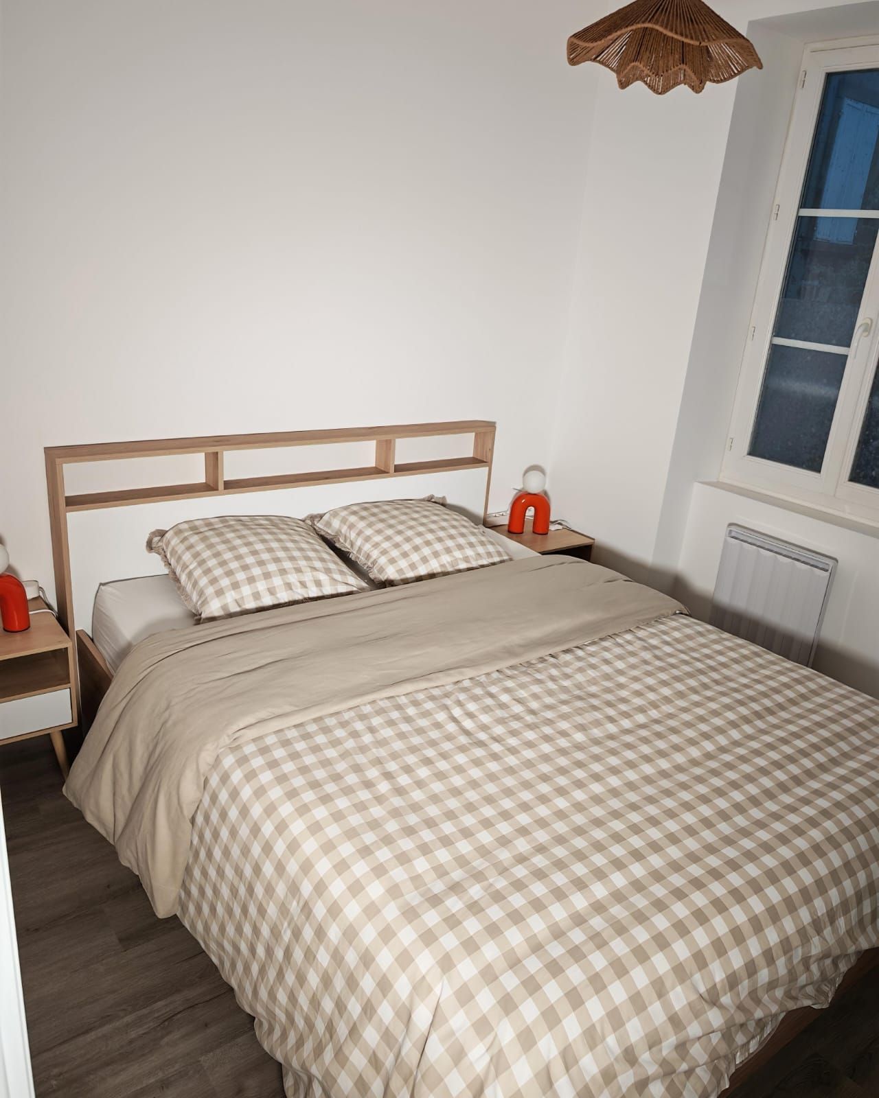 Ein Einzelbett mit Holzseitenverkleidung, einer karierten Tagesdecke, einem Nachttisch mit roter Lampe und einem Fenster mit Jalousien.