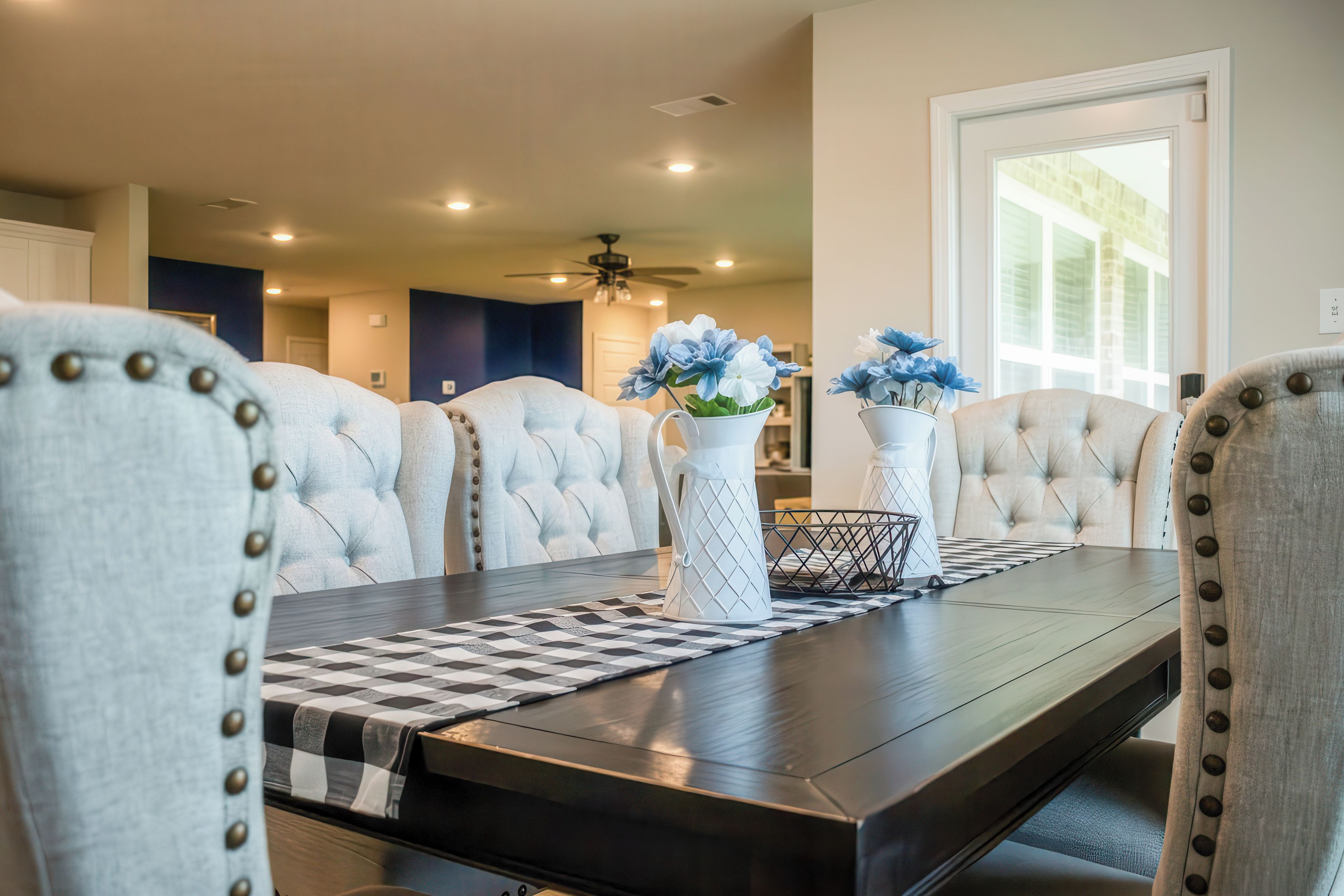 A dining area with a dark wooden table, white upholstered chairs with button details, and two white vases with blue flowers.