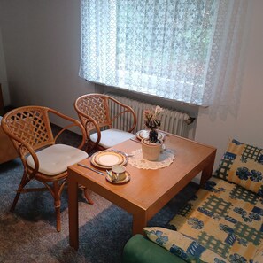 Ein Essbereich mit einem Holztisch, zwei Korbstühlen, einer kleinen Topfpflanze und einem gemusterten Sofa.