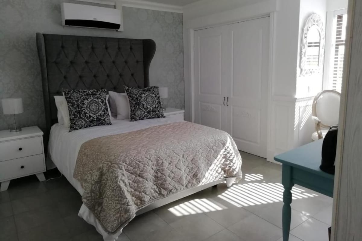 A bedroom with a large bed, a bedside table with a lamp, a wardrobe, and a small table with a chair.