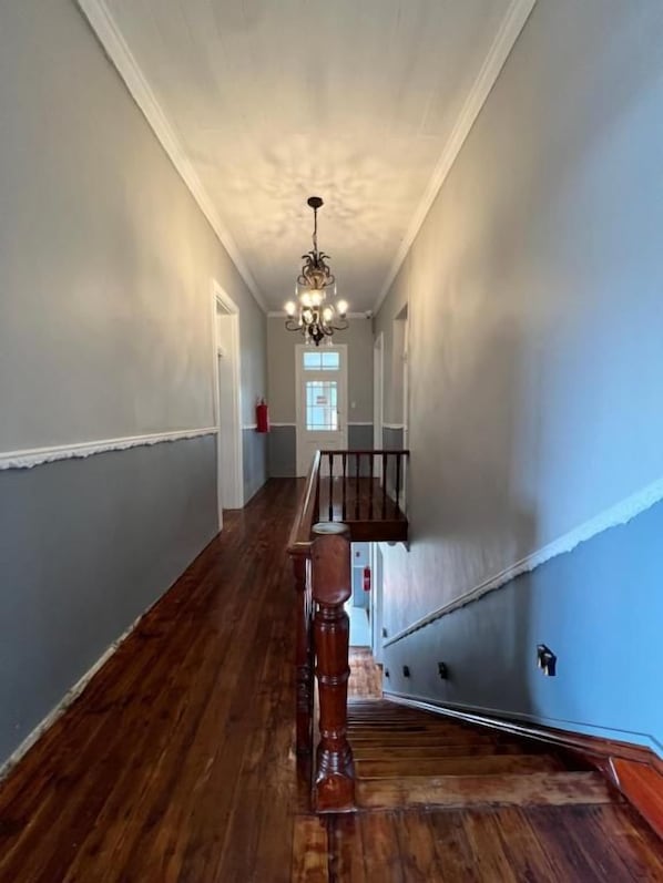 A hallway with a wooden floor, a staircase with a wooden handrail, and a chandelier.