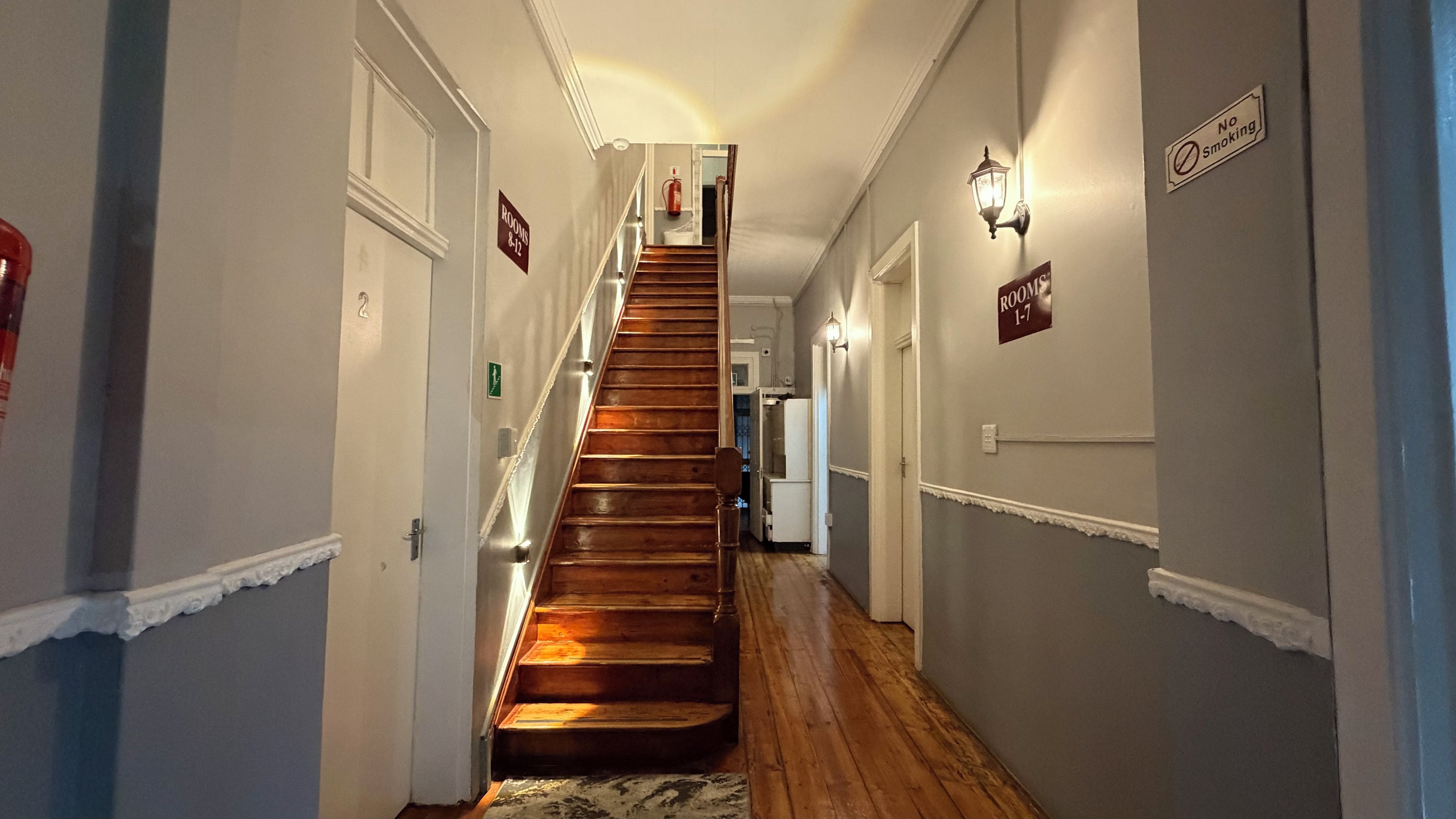 A hallway with a wooden staircase leading to a landing, flanked by doors and signs indicating room numbers.