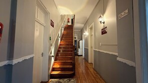 A hallway with a wooden staircase leading to a landing, flanked by doors and signs indicating room numbers.