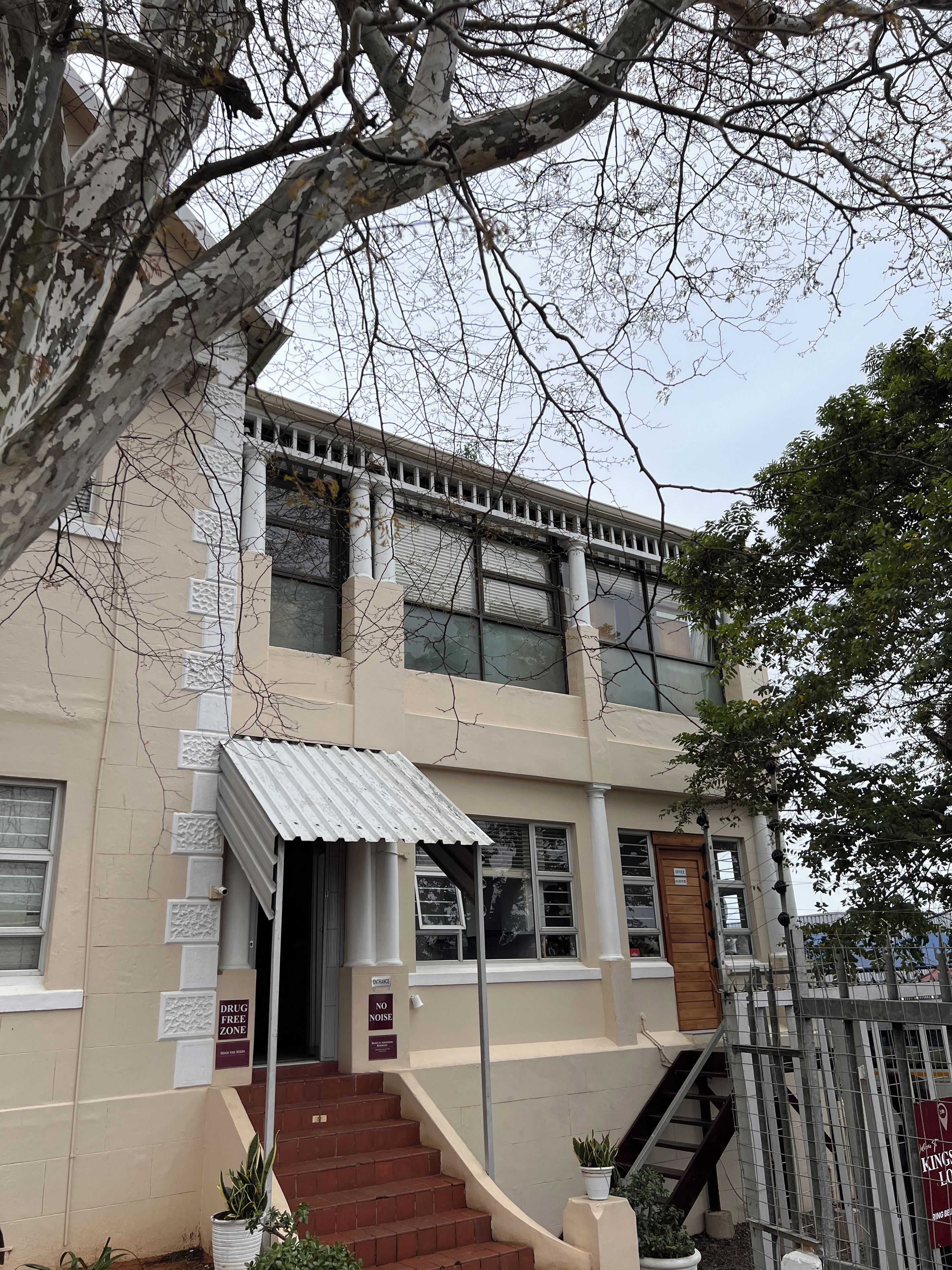 A building with a beige facade, a metal awning over the entrance, and a staircase leading to the entrance. There are signs indicating a 'Drug Free Zone' and 'No Noise' policy.