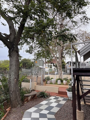 A paved pathway leading to a gated entrance with a metal fence, surrounded by trees and potted plants.