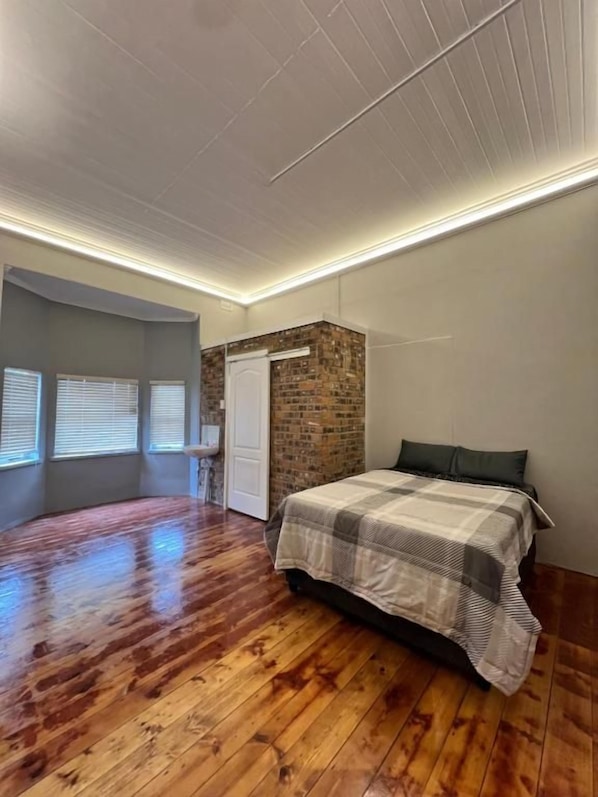 Un dormitorio con piso de madera, cama con colcha a cuadros, pared de ladrillo decorativa y techo blanco.