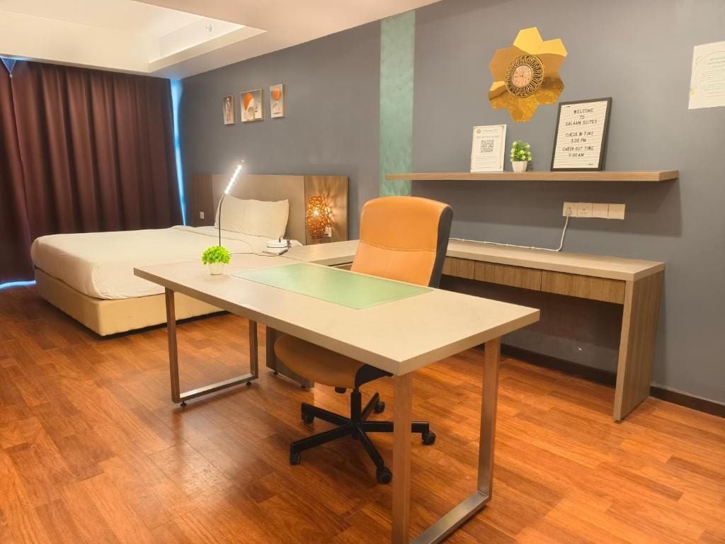 A hotel room with a bed, desk, chair, and a small shelf with a clock and a welcome sign.