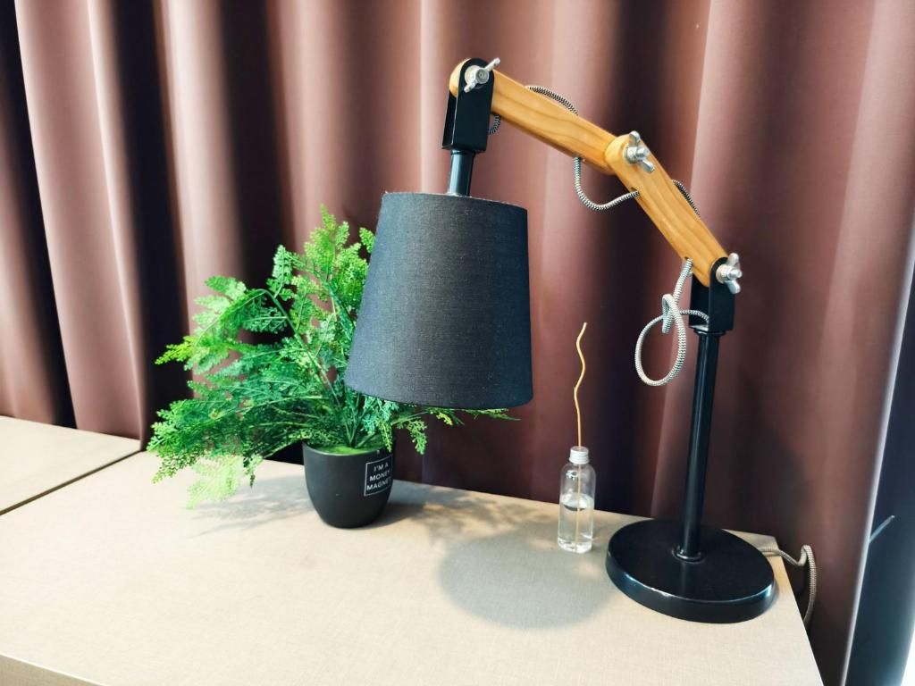 Una lámpara de escritorio con brazo de madera y pantalla negra, una planta en maceta y una pequeña botella sobre una mesa.