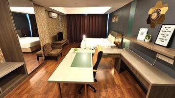 A hotel room with a desk, a bed, a sofa, and a small table. There is a clock on the wall and a sign with check-in times.