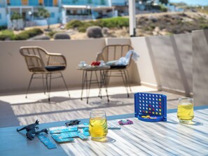 A table with drinks, a toy car, and a game on a sunny terrace with chairs and a building in the background. - Villa Amoudi Private Pool (HERAKLION)
