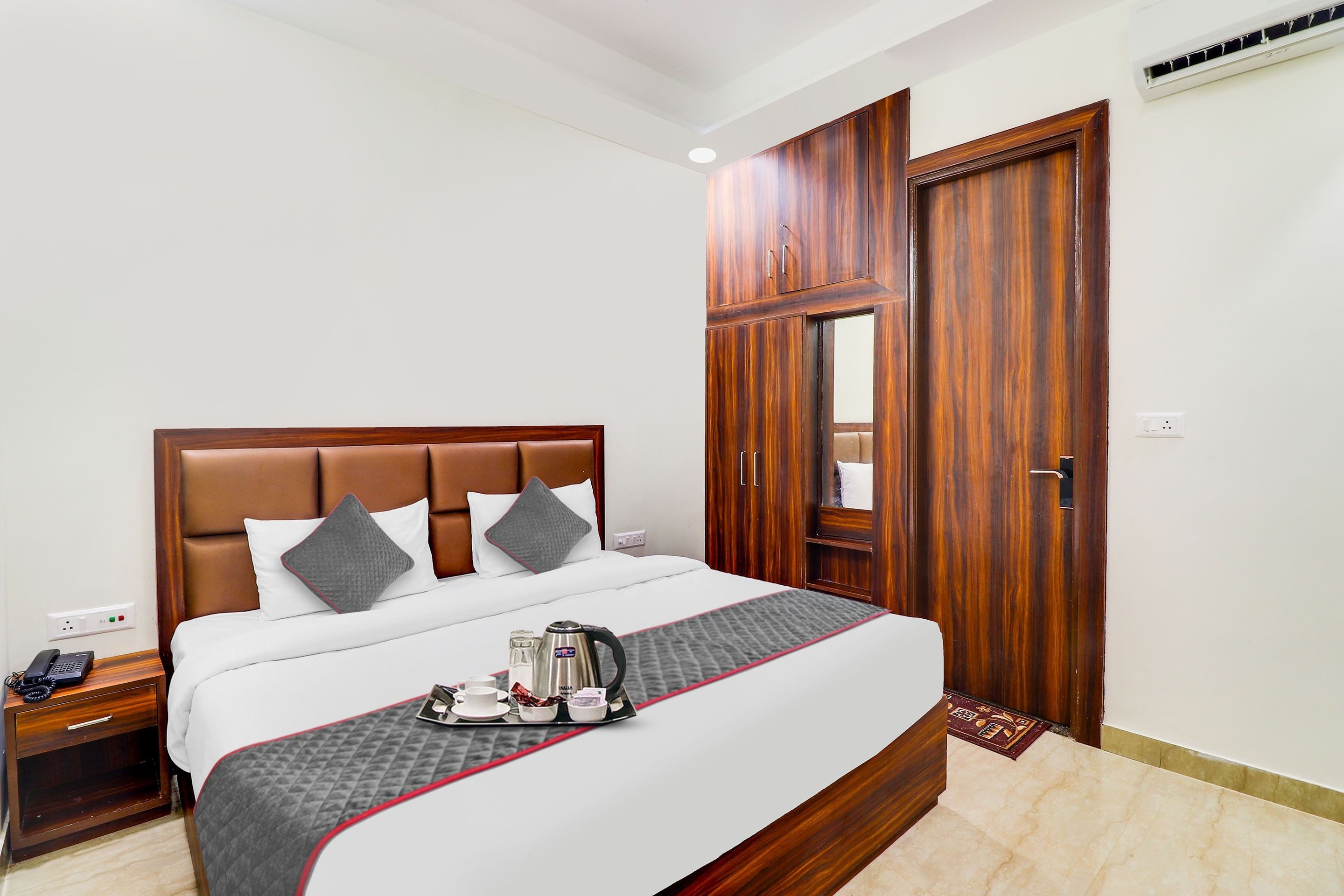 Habitación de hotel con una cama grande, un armario de madera, una mesita de noche con un teléfono y una bandeja con té y tazas.