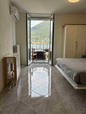 A room with a bed, a table, and a view of a lake and mountains through an open door. - Vrbo Property (Moltrasio)