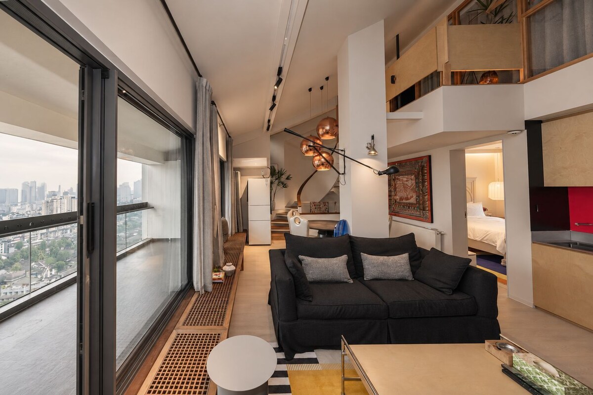 A modern living room with a black sofa, a round table, and a view of the city through large windows.