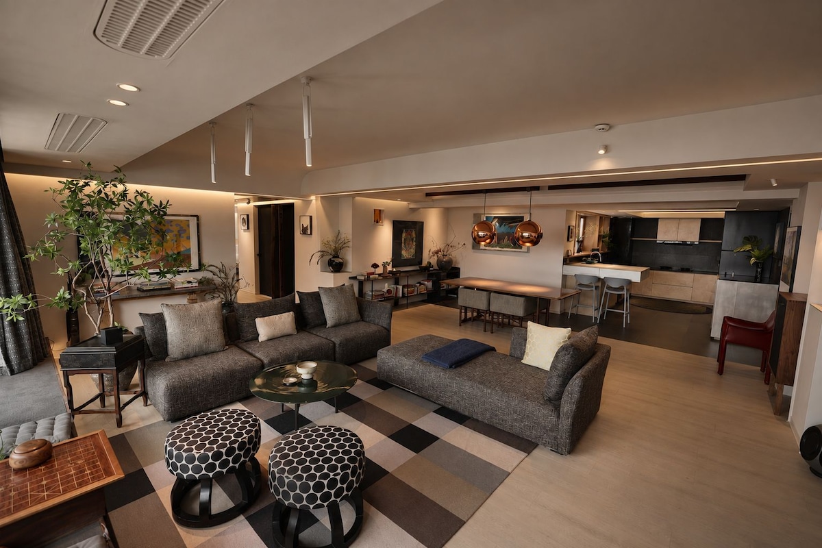 A modern living room with a sofa set, coffee table, and ottomans. There is a dining area with a table and chairs, and a kitchen in the background.