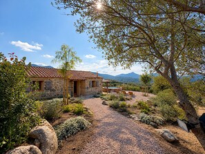 A stone house with a tiled roof, surrounded by a gravel driveway and various plants. - Bergerie Firella (Propriano)
