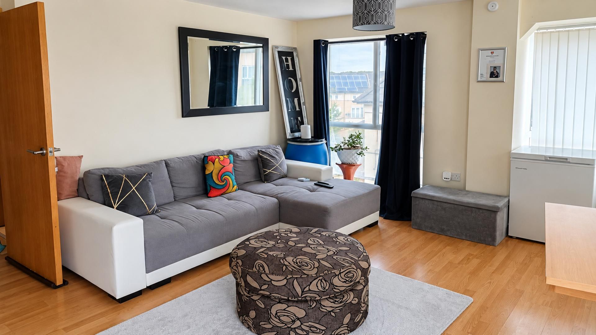 Una sala de estar moderna con un sofá gris, un taburete estampado, una mesita y una ventana con cortinas.