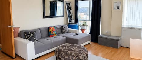A modern living room with a grey sofa, a patterned ottoman, a small table, and a window with curtains.