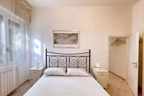 A bedroom with a bed, two bedside tables, a radiator, a window with curtains, a door, and a wall-mounted picture. - UR NEST Toscana - One bedroom apartment inside a gracious villetta (Bologna)