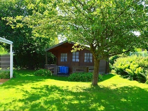 A wooden cabin with a porch, surrounded by lush greenery and a fenced area. - "rungholtstrasse" (Wyk auf Foehr)