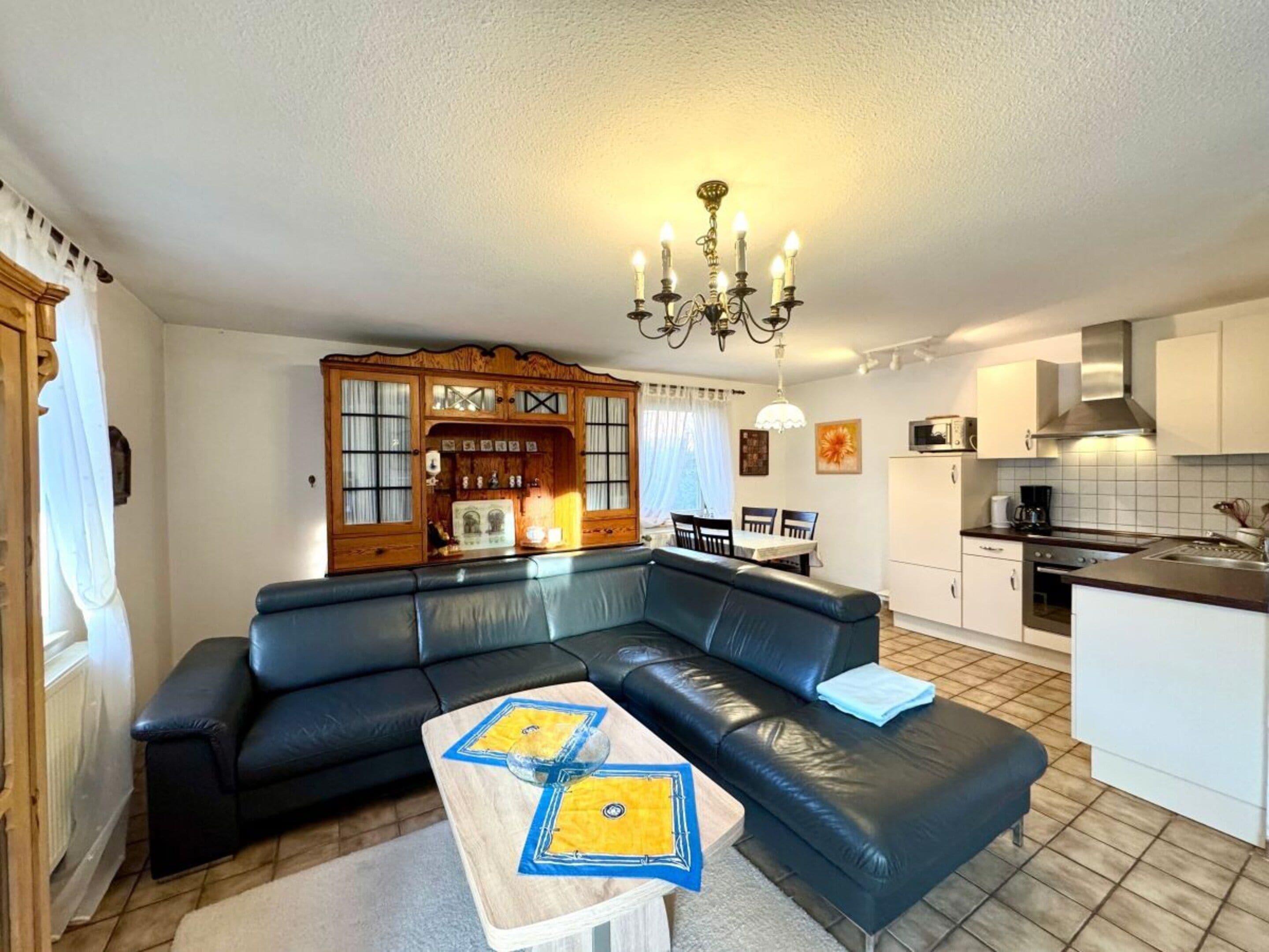 Een moderne woonkamer met een zwarte leren bank, een houten kast met glazen deuren, een eethoek met een tafel en stoelen, en een keuken met witte kasten en een roestvrijstalen afzuigkap.
