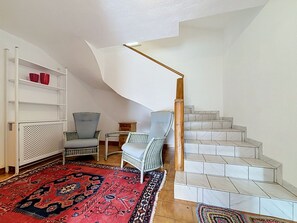 Eine Treppe mit weißen Fliesen, einem rot gemusterten Teppich, einem Korbstuhl und einem weißen Regal mit roten Gegenständen.