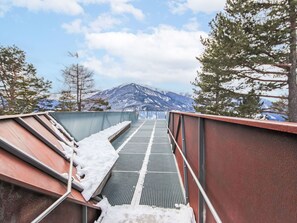Eine Metalltreppe führt zu einer Plattform mit einem Gitterrostboden, umgeben von schneebedeckten Geländern und Bäumen, mit Blick auf einen mit Schnee bedeckten Berg.