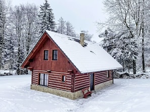 A red wooden cabin with a snow-covered roof, surrounded by snow-laden trees.