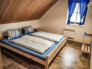 A room with a wooden ceiling, a bed with striped bedding, a small bedside table, a radiator, and a window with curtains.