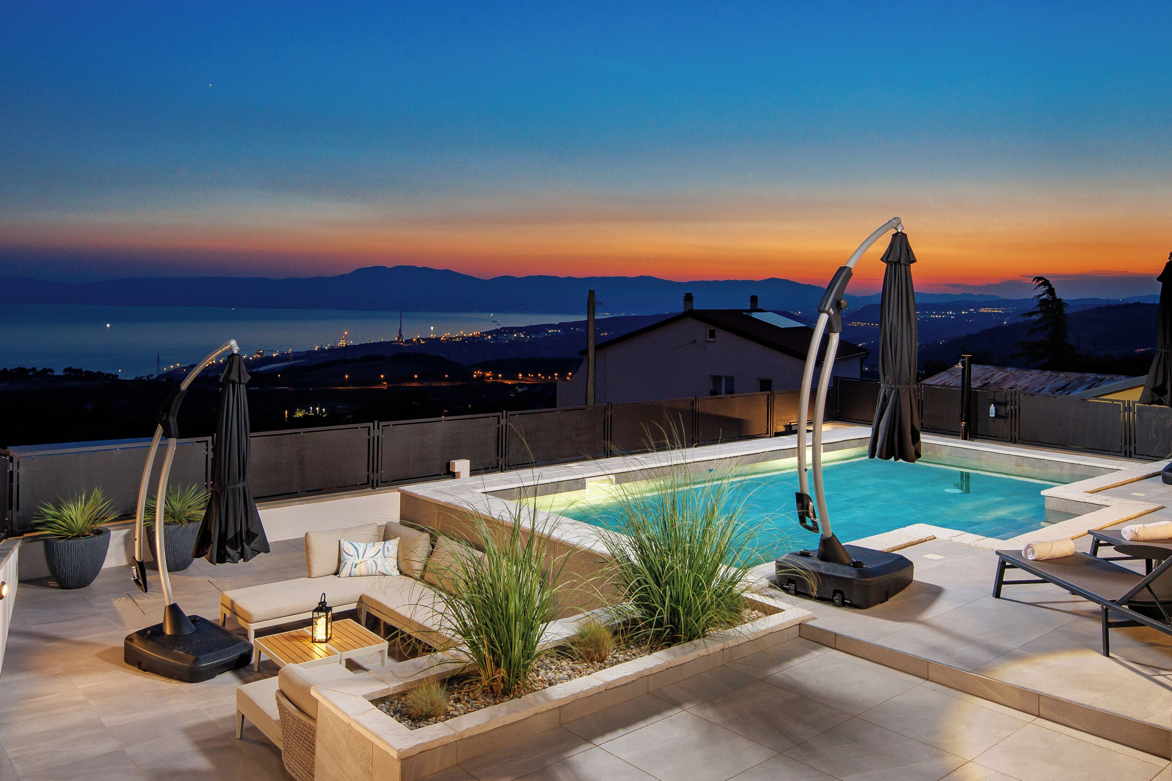A rooftop pool area with lounge chairs, umbrellas, and a view of the city at sunset.