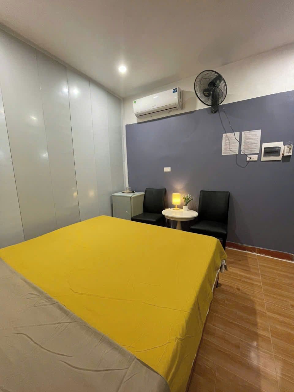 A hotel room with a bed, a small table with a lamp, two chairs, an air conditioning unit, and a wall-mounted fan.