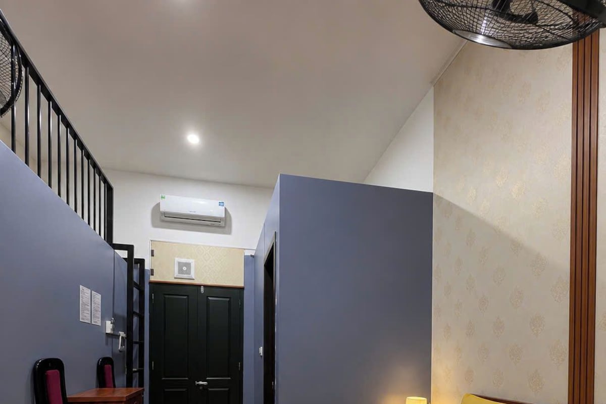 Quarto de hotel com cama, escrivaninha e cadeira, mesinha de cabeceira com abajur e ar-condicionado fixado na parede.