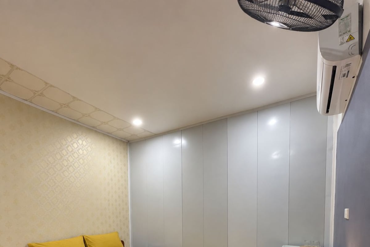 Uma cama de solteiro com edredom amarelo, uma mesa redonda pequena com uma planta, uma cadeira e um ar condicionado fixado na parede.