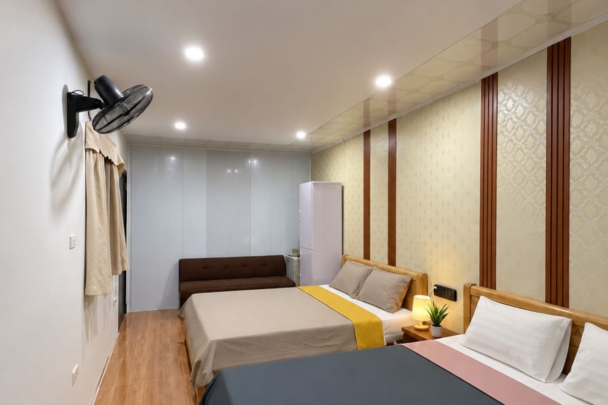 Quarto de hotel com duas camas, um ventilador fixado na parede, uma geladeira e uma mesa de cabeceira com abajur.