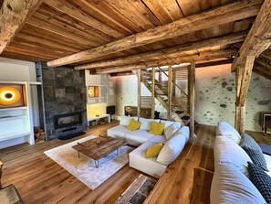Ein rustikales Wohnzimmer mit sichtbaren Holzbalken, einem Steinkamin, einem weißen Sofa mit gelben Kissen und einem Holztisch.