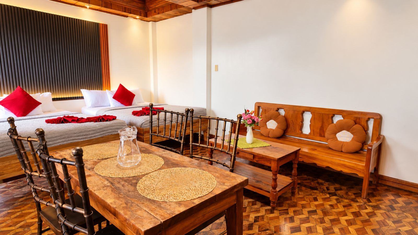 A room with a wooden dining table, chairs, a sofa, and a bed with red pillows.
