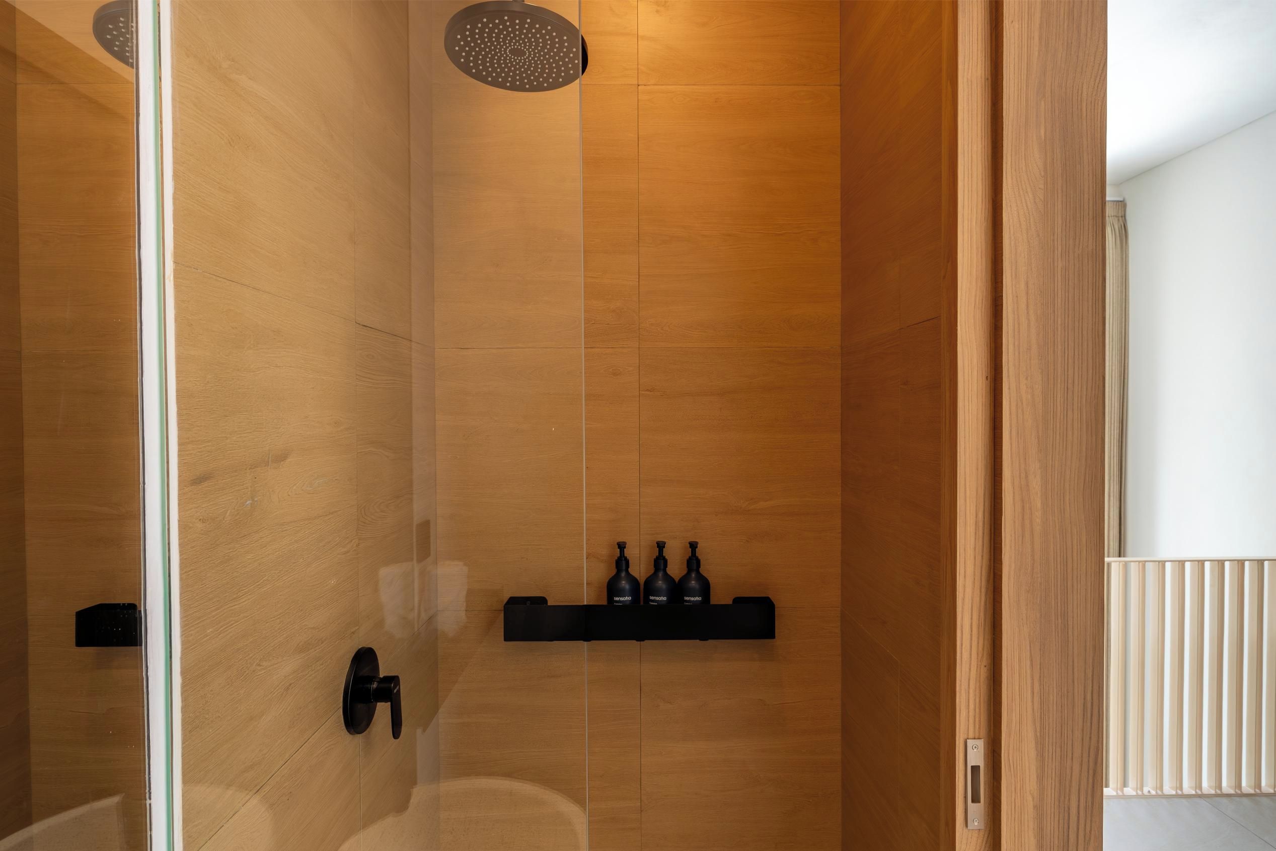 Une salle de bain moderne avec un mur en bois, une cabine de douche en verre et une étagère noire où sont rangés les articles de toilette.