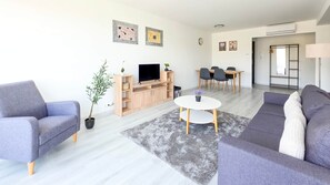 A modern living room with a grey sofa, a coffee table, a TV, and a dining area with chairs and a table. - LB - Balcsikuckó Apartman (Siófok)