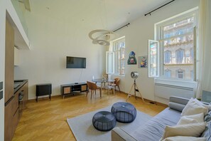 Ein modernes Wohnzimmer mit einer Couch, Hocker, einem Esstisch mit Stühlen, einem an der Wand montierten Fernseher und einem Fenster mit Blick auf Gebäude.