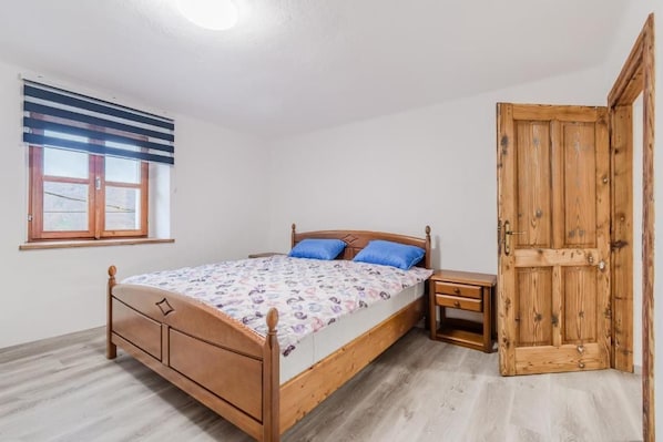 A bedroom with a wooden bed, a nightstand, a window with blinds, and a wooden door.