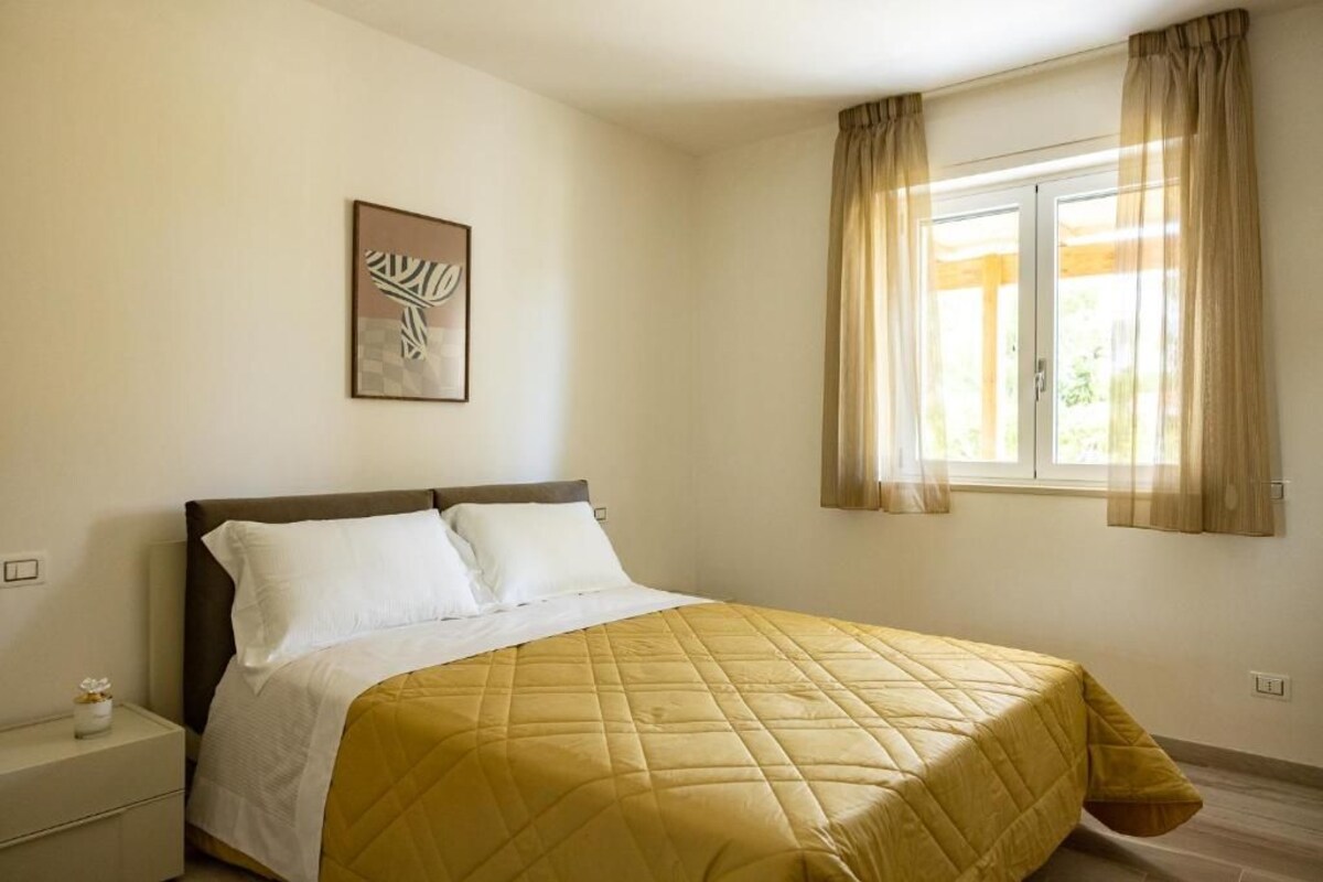 Ein Schlafzimmer mit einem Bett, einem Nachttisch, einem Fenster mit Vorhängen und einem an der Wand hängenden Kunstwerk.