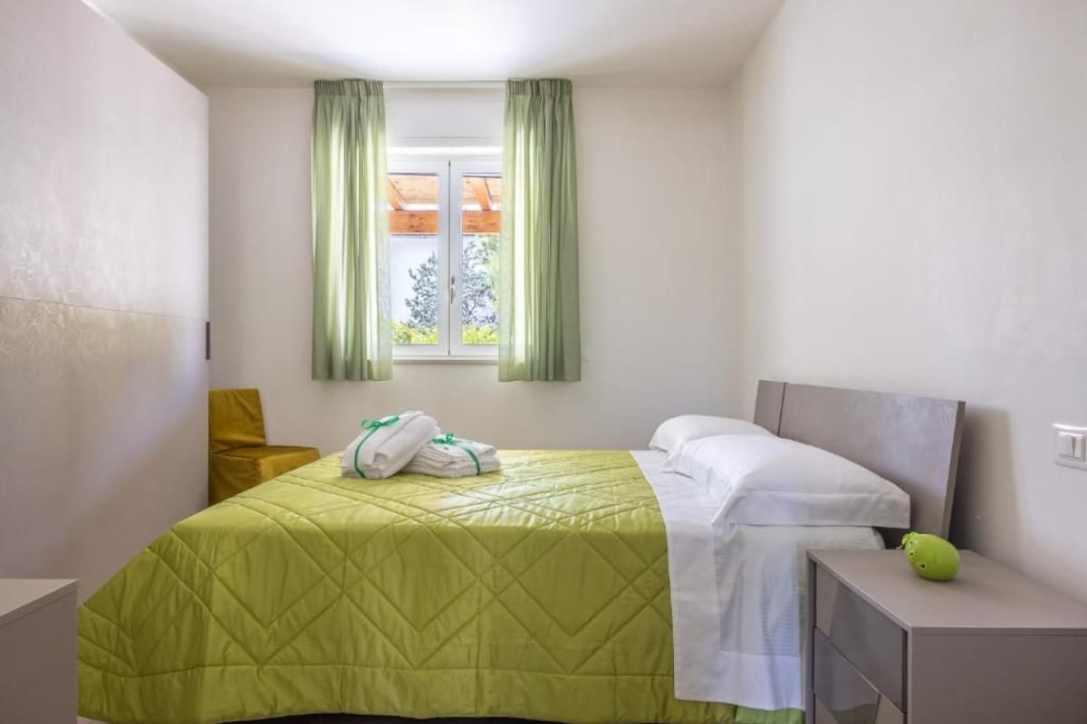 Ein Einzelbett mit einer grünen Tagesdecke, ein Nachttisch mit einem grünen Gegenstand und ein Fenster mit Vorhängen.