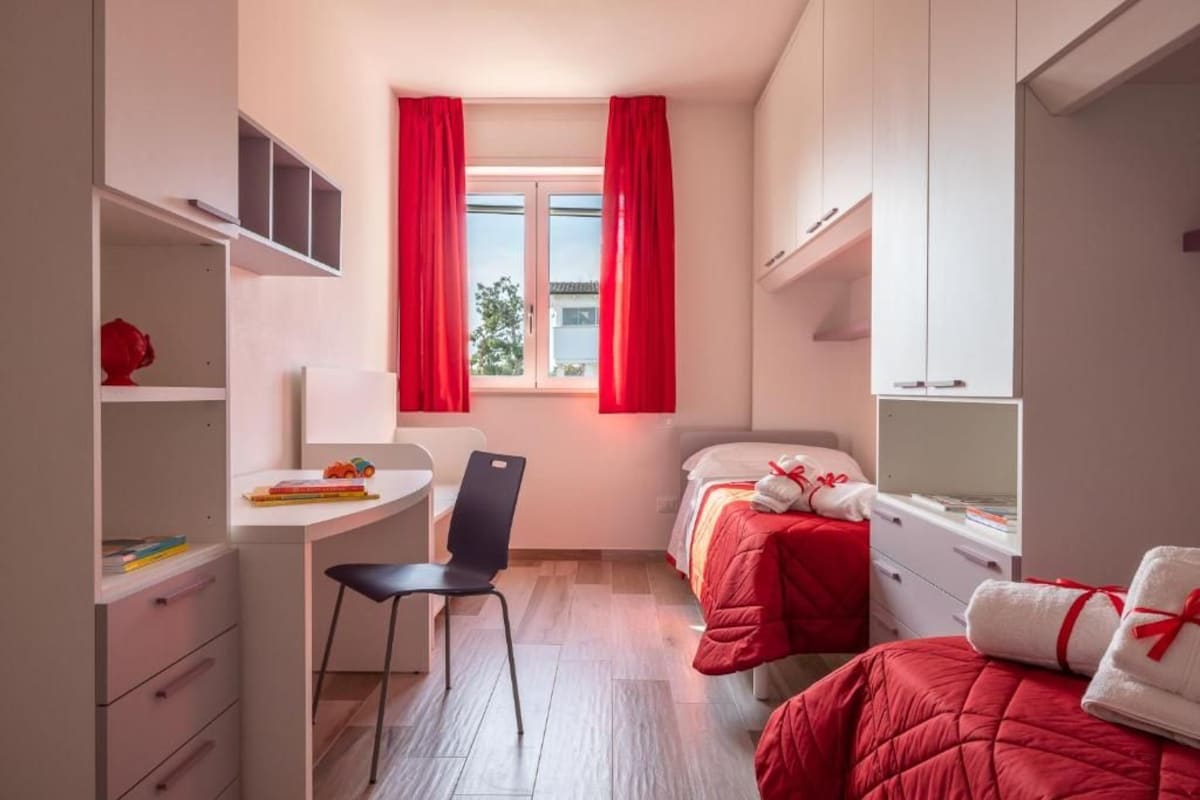 Ein kleines Schlafzimmer mit Schreibtisch, zwei Betten und einem Fenster mit roten Vorhängen.