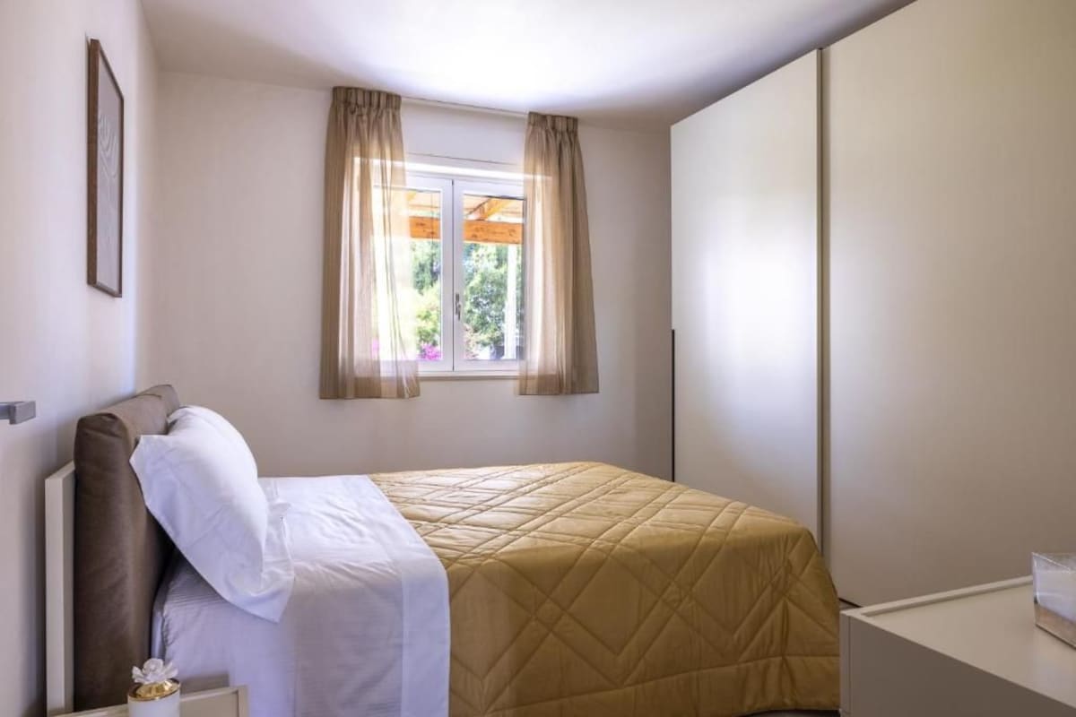 Ein Einzelbett mit gesteppter Bettdecke, ein Nachttisch mit Lampe und ein Fenster mit Vorhängen.