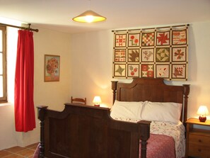 Quarto com cama de madeira, colcha pendurada na parede, cortina vermelha, pintura e abajur.