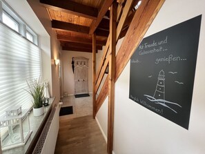 Passadís amb bigues de fusta, una planta en test i una porta blanca, i un cartell de benvinguda amb una il·lustració d'un far.