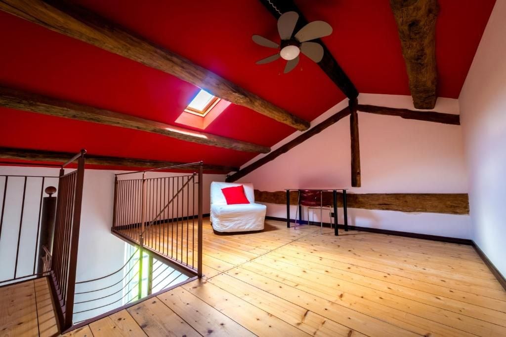 Una habitación con techo inclinado, vigas de madera, una silla blanca con cojín rojo y un ventilador de techo.