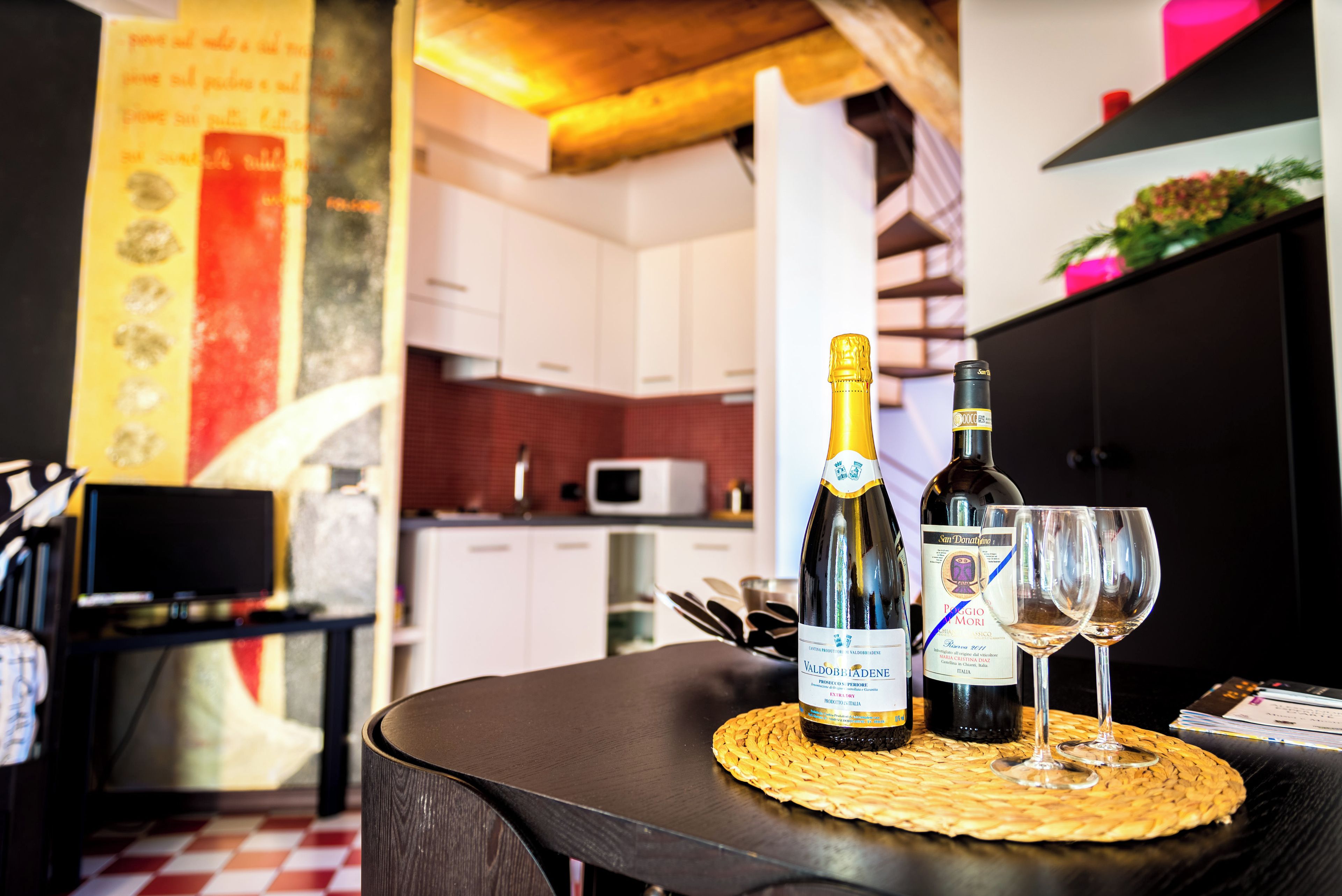 Un tavolo con una bottiglia di vino e due bicchieri, un sottopiatti intrecciato e una cucina sullo sfondo.